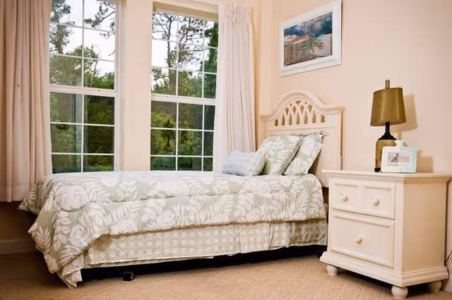 A cozy bedroom with a single bed covered in a floral patterned comforter and pillows. The bed is positioned next to large windows with white curtains, allowing natural light to brighten the room. Beside the bed is a white nightstand with two drawers, holding a lamp and a small framed photo. A framed picture hangs on the wall above the bed.