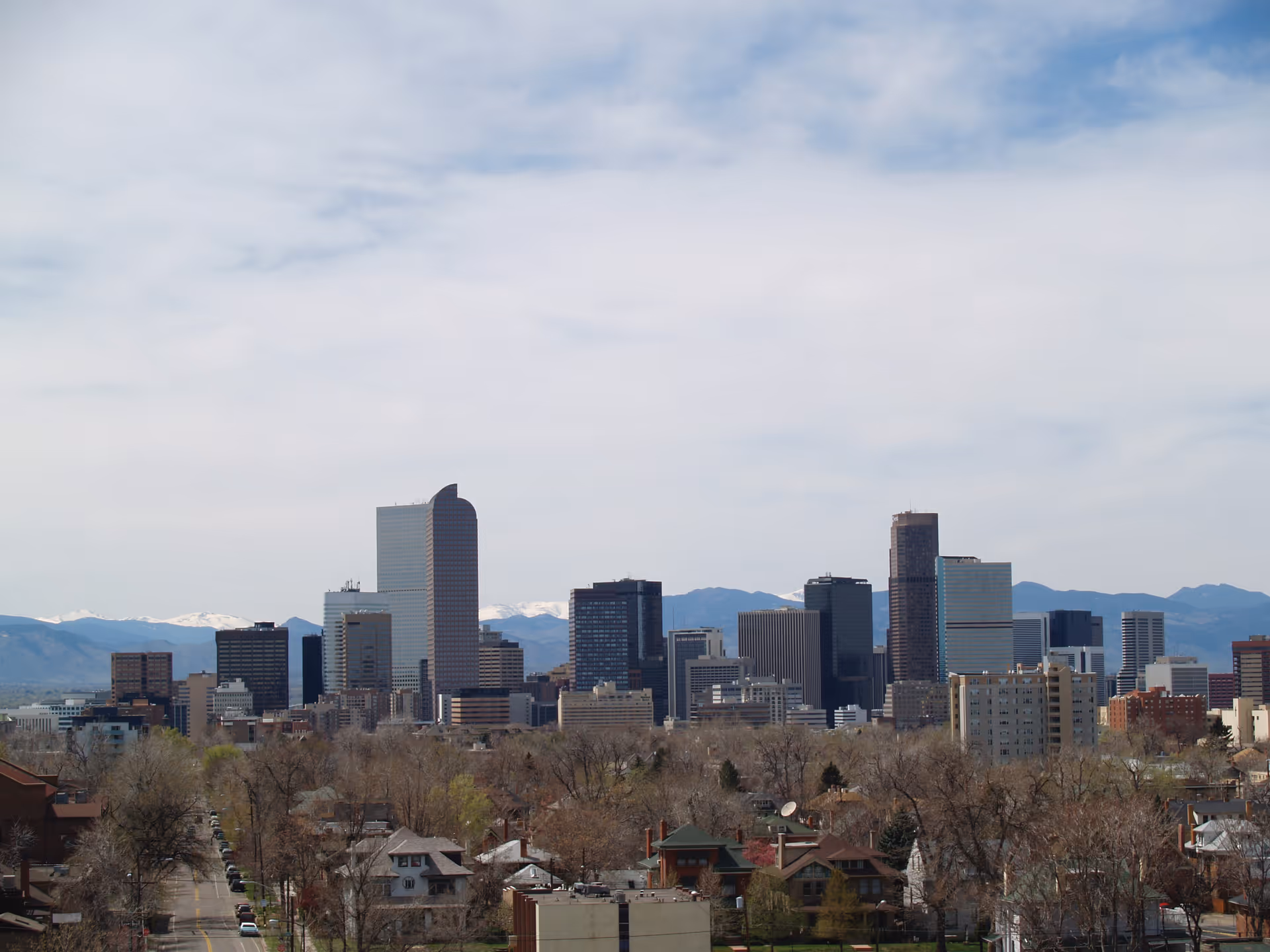 View of a city skyline with tall buildings and mountains in the background under a partly cloudy sky. In the foreground, there are residential houses and trees.