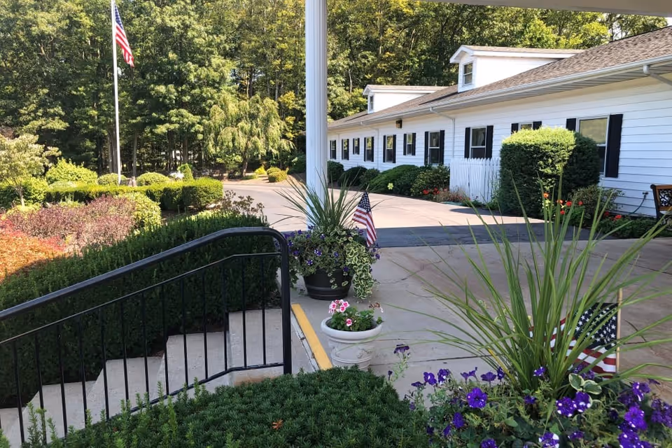 Outdoor view of a senior living facility with a white building featuring black shutters, surrounded by well-maintained bushes, flowers, and American flags. There is a concrete walkway with a black metal railing and a flagpole with an American flag in the background, set against a backdrop of trees.