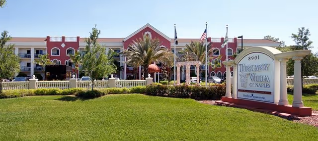 Front exterior view of Tuscany Villa of Naples, featuring a large green lawn, palm trees, a white and red entrance sign with the facility name, and a multi-story building with balconies in the background under a clear blue sky.