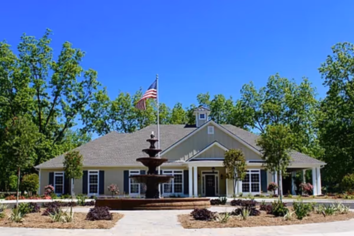 Front exterior view of a single-story building with a gray roof and beige siding, surrounded by trees and landscaping. There is a multi-tiered fountain in the center of a circular driveway, and an American flag flying on a flagpole behind the fountain.