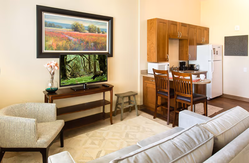A cozy living area with a beige sofa and a striped armchair facing a wooden TV stand with a flat-screen TV displaying a forest scene. Above the TV is a framed landscape painting of a field with trees and a blue sky. To the right is a small kitchen area with wooden cabinets, a white refrigerator, microwave, coffee maker, and a small dining table with two wooden chairs. The room has light-colored walls and a patterned beige rug on the floor.