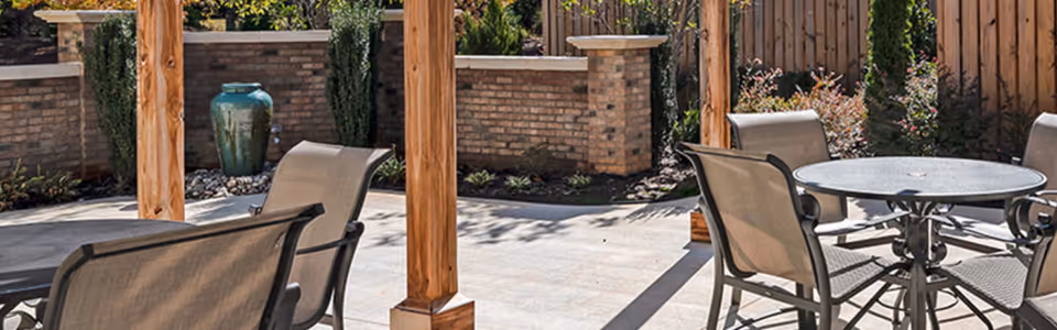 Outdoor patio area with a round metal table and four mesh chairs under a wooden pergola. The background features a brick wall, wooden fence, plants, and a large decorative green vase.