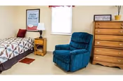 A small bedroom with a single bed and quilted bedspread, a nightstand with lamp, a teal recliner chair, and a wooden dresser under a window.