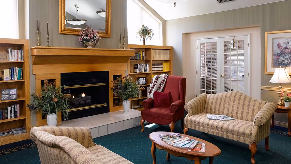 A cozy living room area with a lit fireplace surrounded by wooden shelves filled with books and decorative items. There is a red armchair with a blanket draped over it, a striped beige loveseat, and a matching armchair. A wooden coffee table with magazines is in the center. The room has green carpet, a large mirror above the fireplace, a table lamp on a side table, and French doors with blinds.