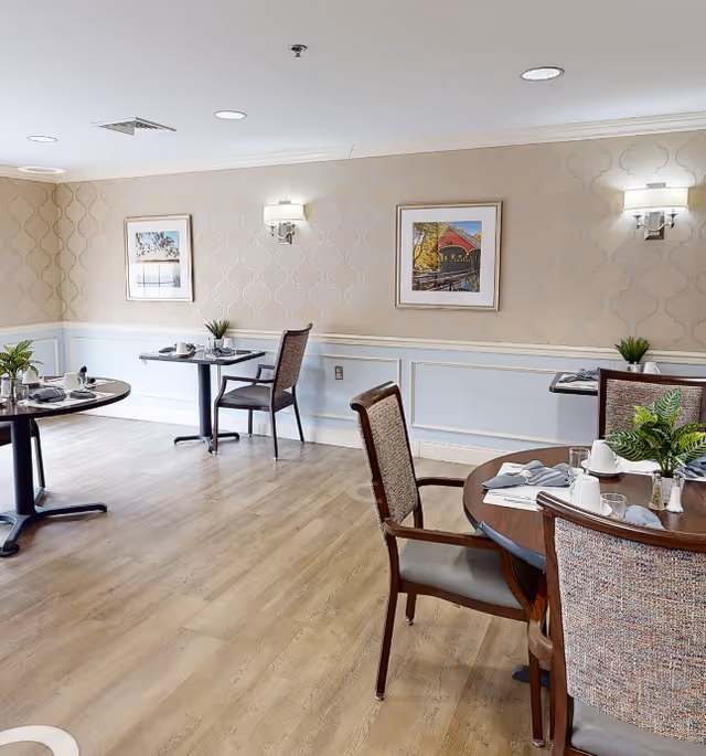 A bright dining room with wooden floors and light-colored walls featuring a subtle patterned wallpaper. There are several round tables set with white cups, plates, napkins, and silverware. Each table has chairs with cushioned seats and backs, and small green plants as centerpieces. Two framed pictures hang on the wall, and there are wall-mounted light fixtures providing soft lighting.