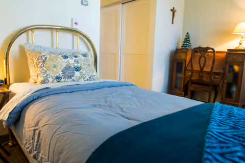 A cozy bedroom with a single brass-headboard bed made with blue bedding, a wooden desk and chair with a lamp, and sliding closet doors.