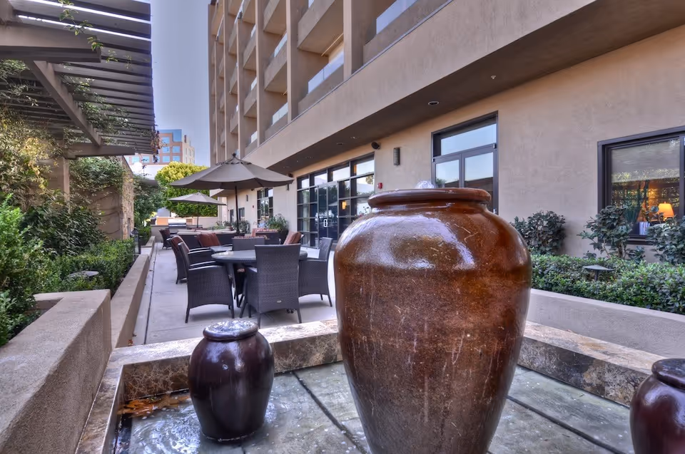 Outdoor patio area at a senior living facility with large ceramic water fountains in the foreground, surrounded by seating with tables and umbrellas, adjacent to a building with large windows and greenery along the sides.