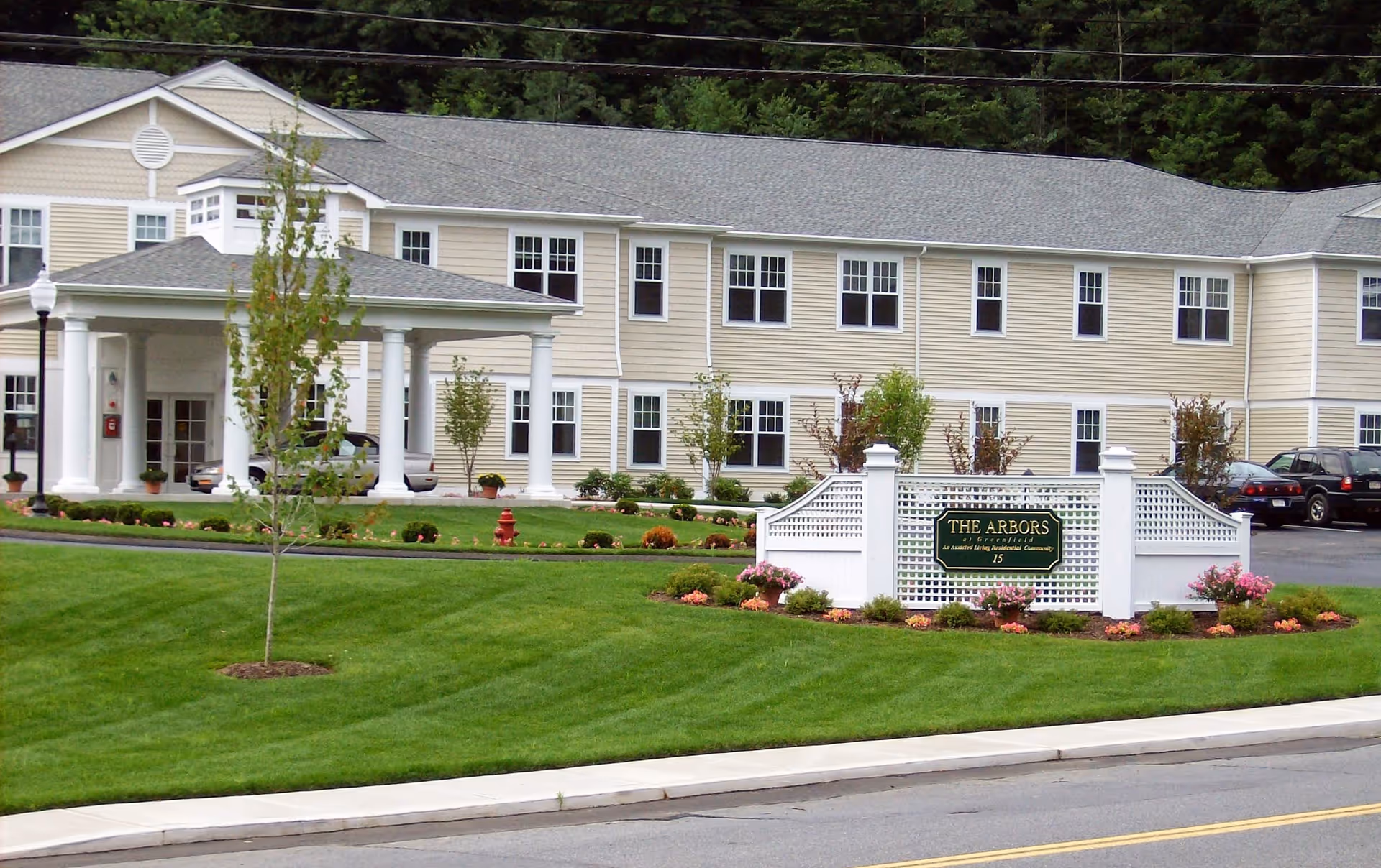 Front exterior of a beige two-story assisted living building with a columned entrance, manicured lawn, and a white sign reading "The Arbors."