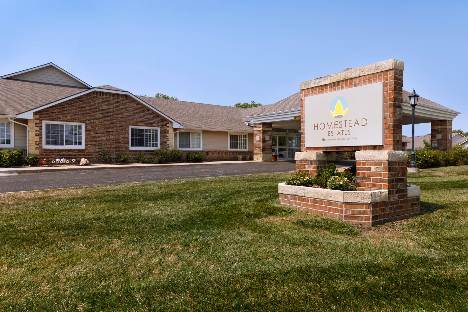 Exterior view of Homestead Estates of Wichita, showing a brick building with multiple windows and a covered entrance. In the foreground, there is a large brick sign with the Homestead Estates logo and name, surrounded by a grassy lawn under a clear blue sky.