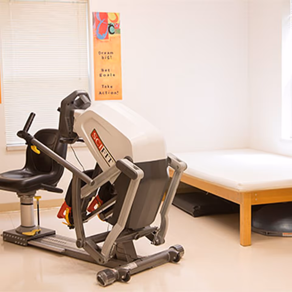 A small therapy room with a recumbent exercise machine and a treatment table against a plain wall.