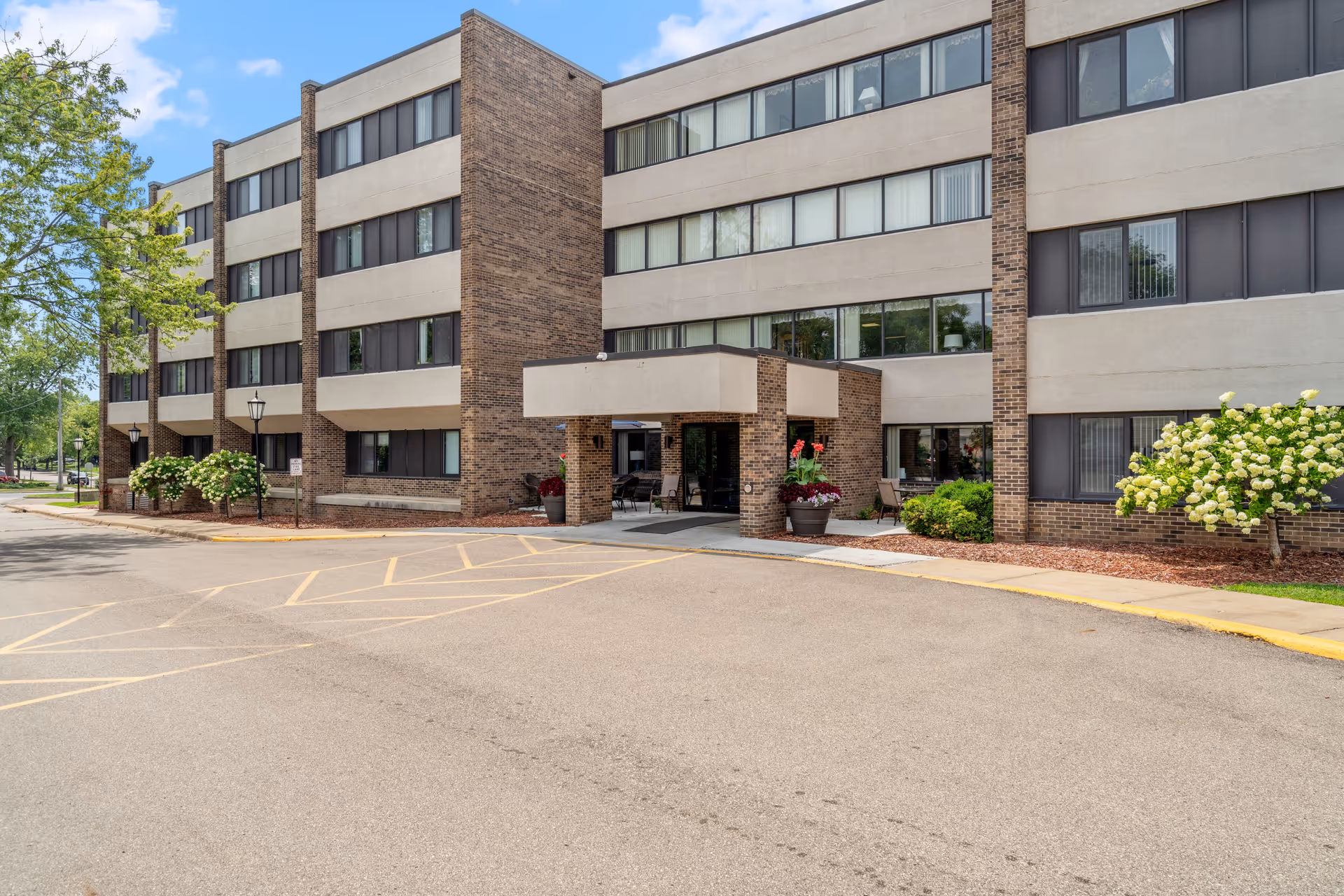 Exterior view of a multi-story brick and concrete building with large windows, an entrance canopy, and landscaped areas with bushes and trees under a partly cloudy sky.