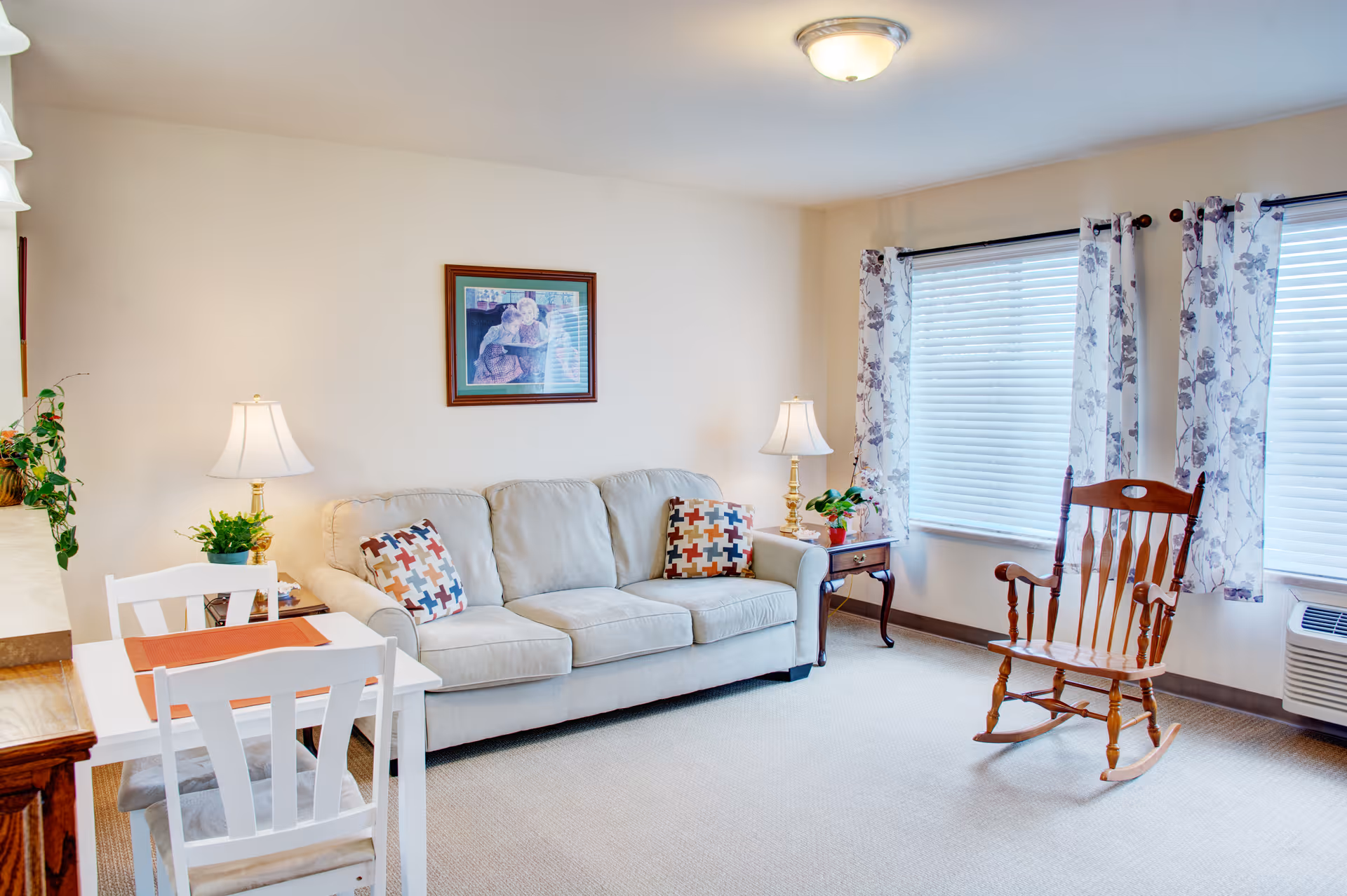 Bright furnished living room with a beige sofa, patterned pillows, a wooden rocking chair, side tables with lamps, a small dining table, and windows with floral curtains.
