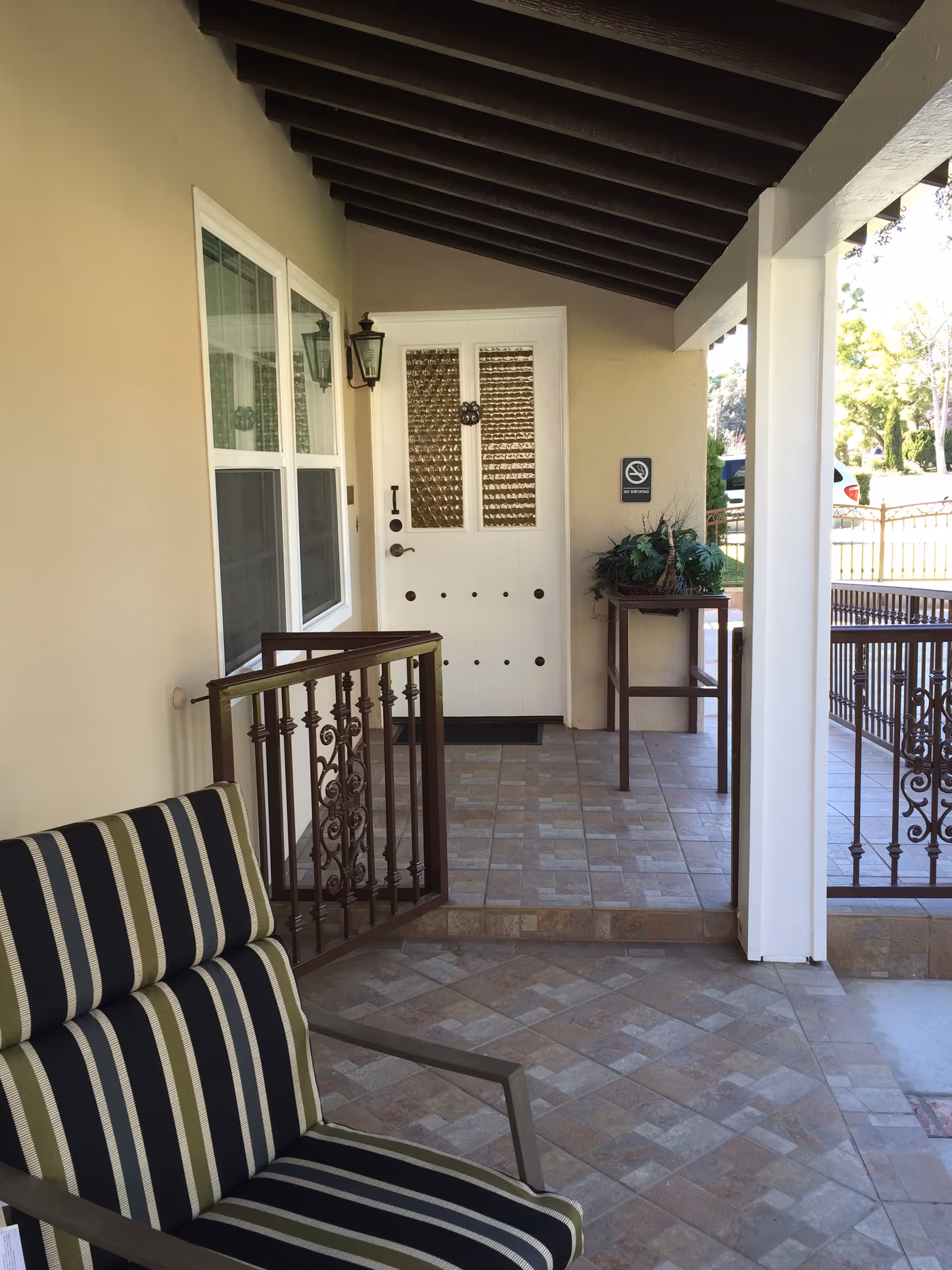 Covered porch area with a striped cushioned chair in the foreground, a white door with textured glass panels, a small table with a plant, and decorative metal railings. The porch has tiled flooring and a wooden beam ceiling.