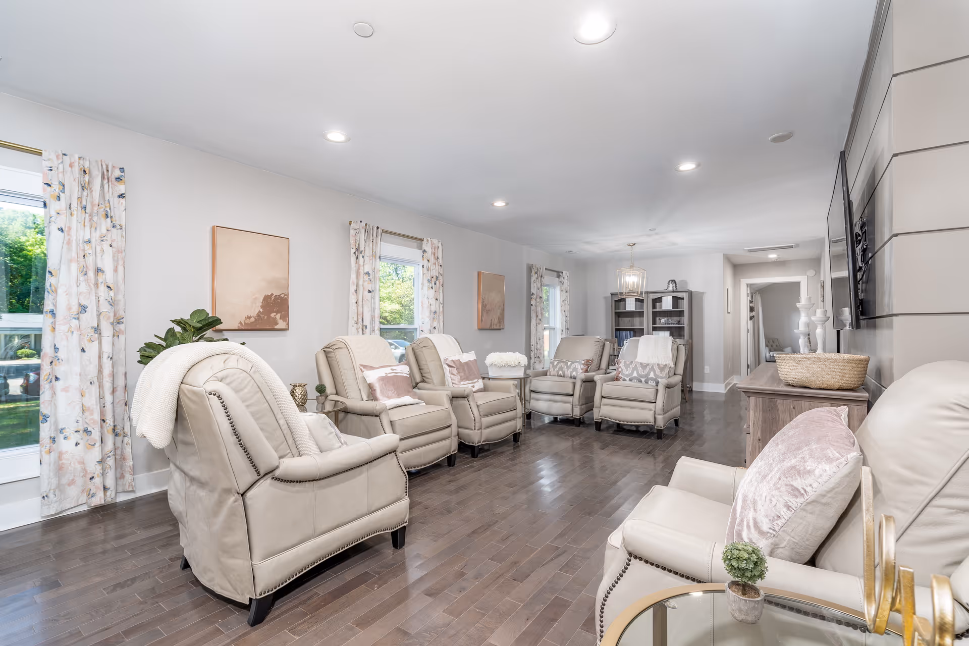 A bright and spacious living room with six beige recliner chairs arranged in a semi-circle facing a wall-mounted flat-screen TV. The room has hardwood floors, light-colored walls, and three windows with floral curtains letting in natural light. There are decorative pillows on the chairs, a small round glass table with a plant, and a wooden cabinet with decorative items. The room has recessed ceiling lights and a cozy, inviting atmosphere.