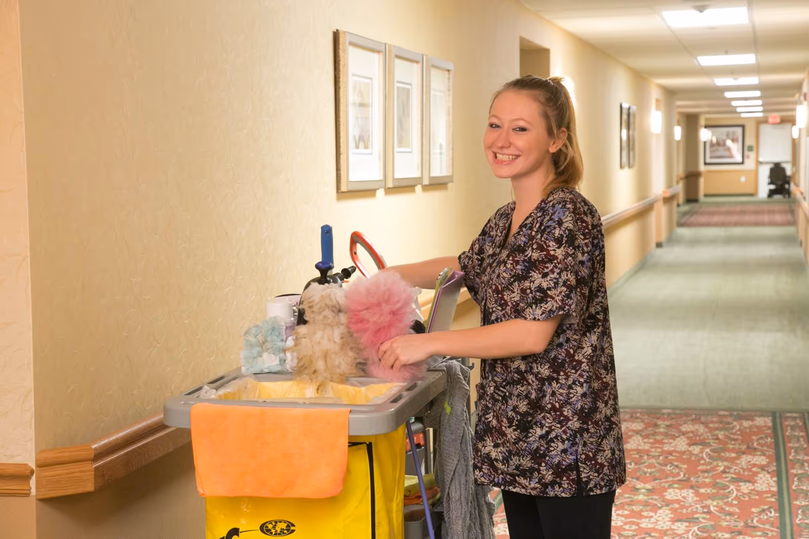 A smiling woman standing in a hallway next to a cleaning cart filled with cleaning supplies and dusters. The hallway has beige walls, framed pictures, and a patterned carpet.