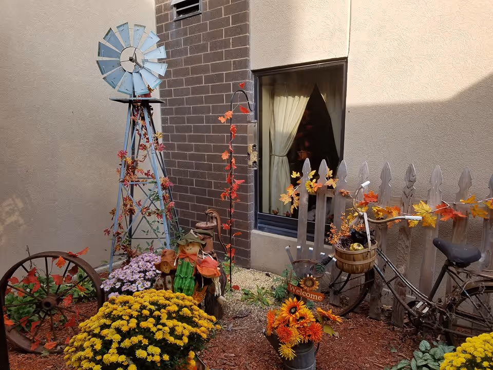 A small outdoor garden area with a decorative blue windmill, colorful flowers including yellow and purple blooms, a rustic wooden fence adorned with autumn leaves, a vintage bicycle with a basket filled with fall decorations, and a 'Welcome' sign. The garden is situated next to a building with a window and brick wall.