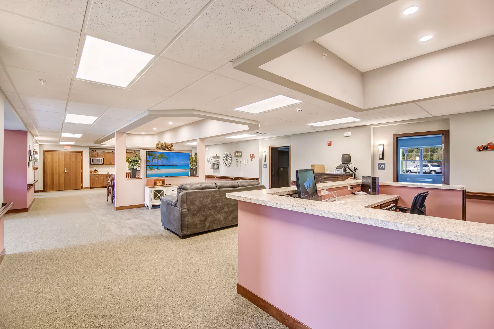 Interior view of a senior living facility reception area with a pink front desk, computer monitors, and chairs. Behind the reception is a common area with a gray couch facing a large flat-screen TV displaying a beach scene. The space is well-lit with ceiling lights and has beige carpeting and light-colored walls. In the background, there is a hallway leading to other rooms and a kitchen area with wooden cabinets and a microwave.