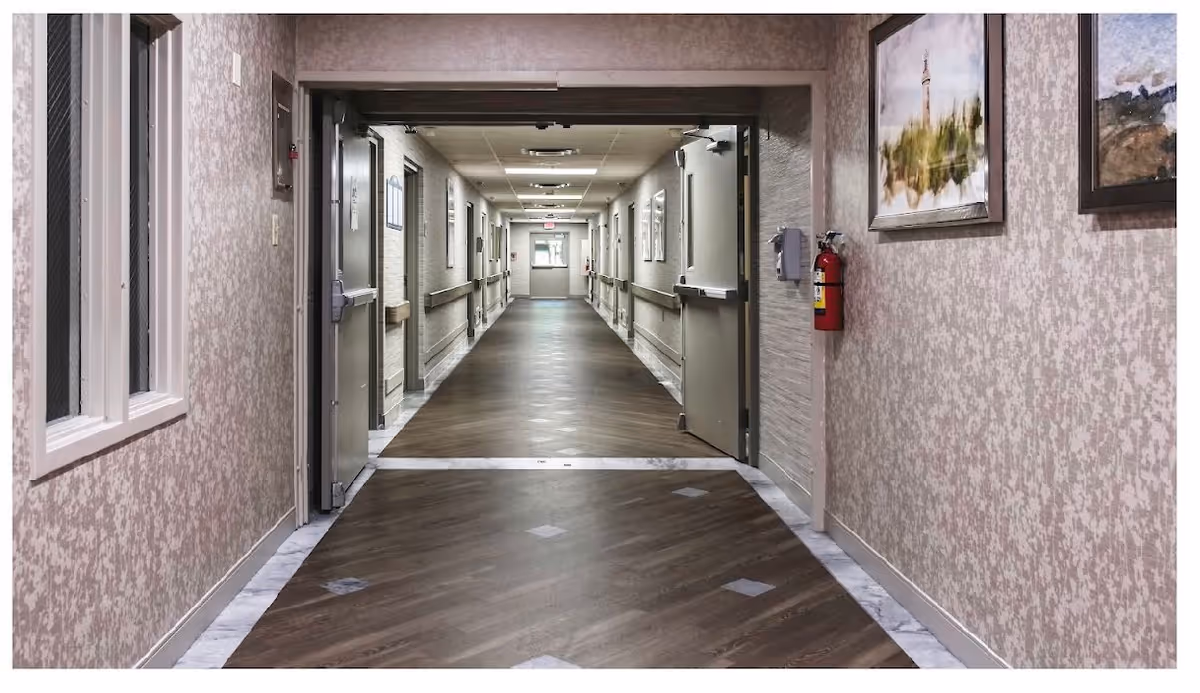 A long, empty hallway in a nursing and rehabilitation facility with closed doors on both sides, handrails along the walls, framed artwork, and a fire extinguisher mounted on the right wall.