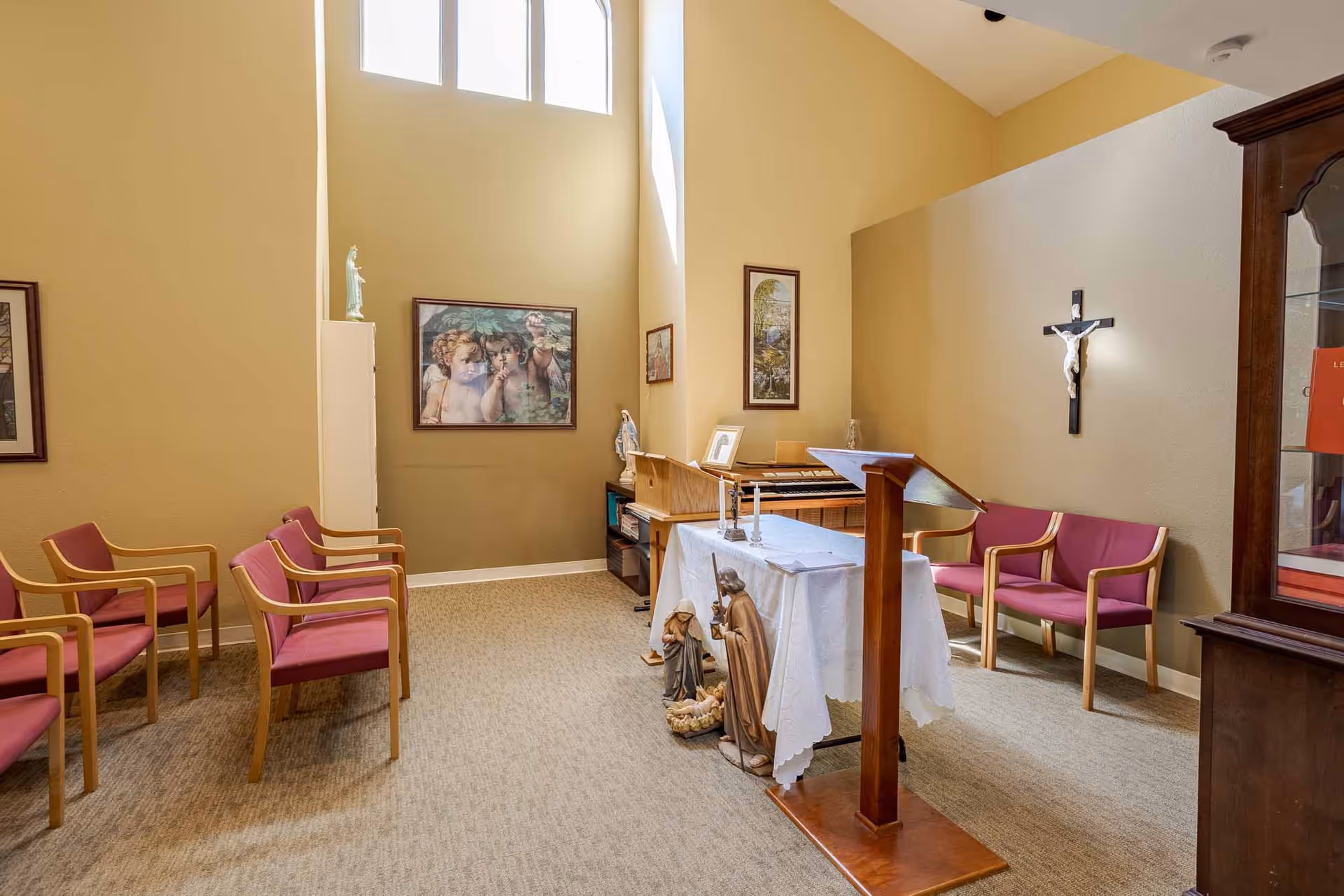 A small chapel room with beige walls and carpeted floor. There are several wooden chairs with red cushions arranged along the walls. A wooden podium stands in the center with a white cloth-covered altar table behind it, adorned with religious statues and candles. A crucifix is mounted on the wall to the right, and framed religious artwork decorates the walls. Tall windows near the ceiling allow natural light to enter the room.