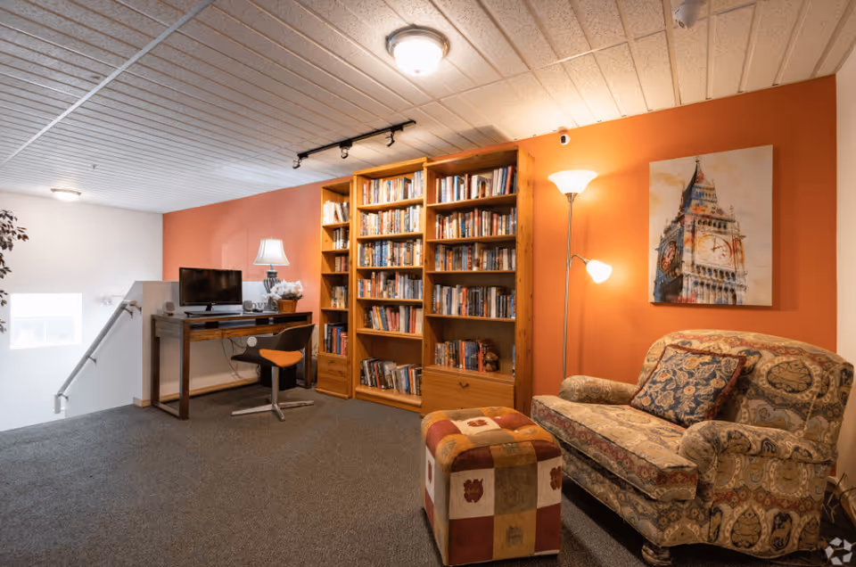 Cozy interior room with an orange accent wall featuring a large wooden bookshelf filled with books, a patterned loveseat with decorative pillows, a colorful ottoman, a floor lamp, and a desk with a chair and a small TV. A painting of Big Ben hangs on the wall above the loveseat.