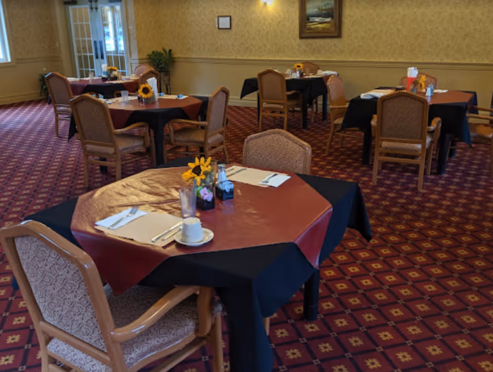 Dining room with multiple tables covered with black and red tablecloths, each set with placemats, utensils, cups, and small flower arrangements. The room has patterned carpet, beige walls with wainscoting, and framed artwork on the walls.