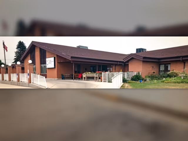 Exterior view of Good Samaritan Society - Bottineau, a single-story brick building with a sloped roof. There is a white fence along the front entrance, a flagpole with an American flag, and some greenery and flowers near the building.