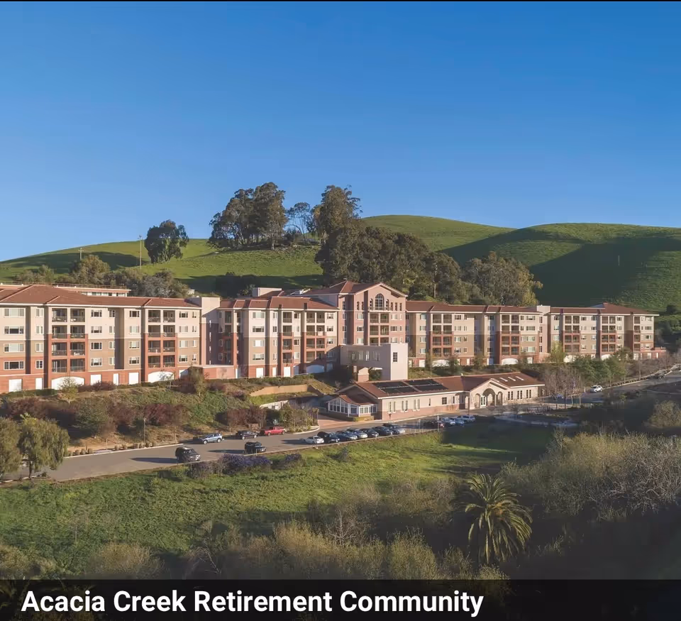 A multi-story retirement community building set against green rolling hills under a clear blue sky.