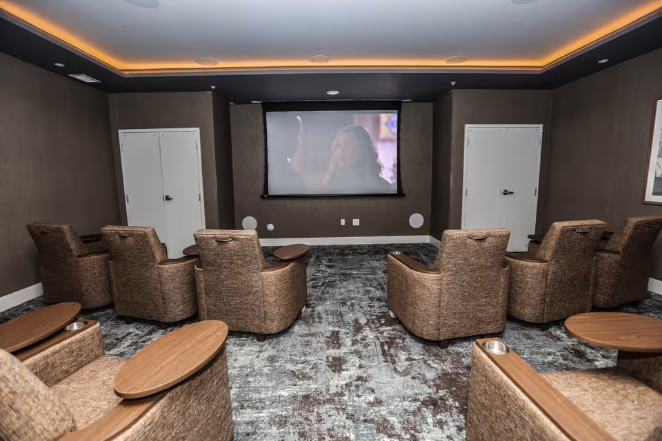 A small theater room with eight brown upholstered recliner chairs arranged in two rows facing a large projection screen on the front wall. The room has dark walls, a patterned carpet, recessed lighting with a warm glow around the ceiling perimeter, and two white double-door closets on either side of the screen.