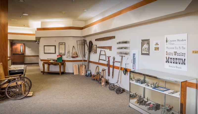 Interior hallway of Village Concepts of Sedro-Woolley - Country Meadow Village featuring a display of vintage tools and artifacts along the wall, a glass display case with small items, a wooden table with framed pictures, and a sign indicating the Sedro-Woolley Museum Satellite Branch.