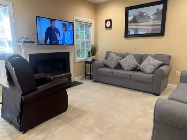 A cozy living room with beige walls and carpet. There is a gray sofa with four patterned cushions, a dark recliner chair, and a second gray sofa partially visible. A flat-screen TV is mounted above a black fireplace, showing a scene with two people. A small side table with a plant and a clock on the wall are also visible. A framed black and white picture hangs above the sofa.