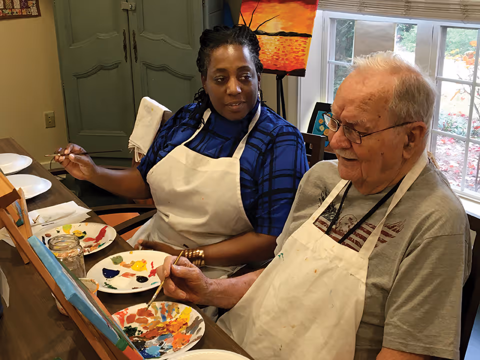 Two people sitting at a table painting on canvases. Both are wearing aprons and holding paintbrushes. There are paint palettes with various colors on the table. A window with a view of greenery is in the background.