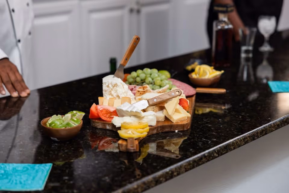 A close-up view of a kitchen counter with a wooden cutting board displaying various cheeses, grapes, tomato slices, and crackers. Two cheese knives are stuck into the cheeses. There are small bowls with lime wedges and lemon slices on the counter, and a person wearing a white chef's coat is partially visible on the left side.