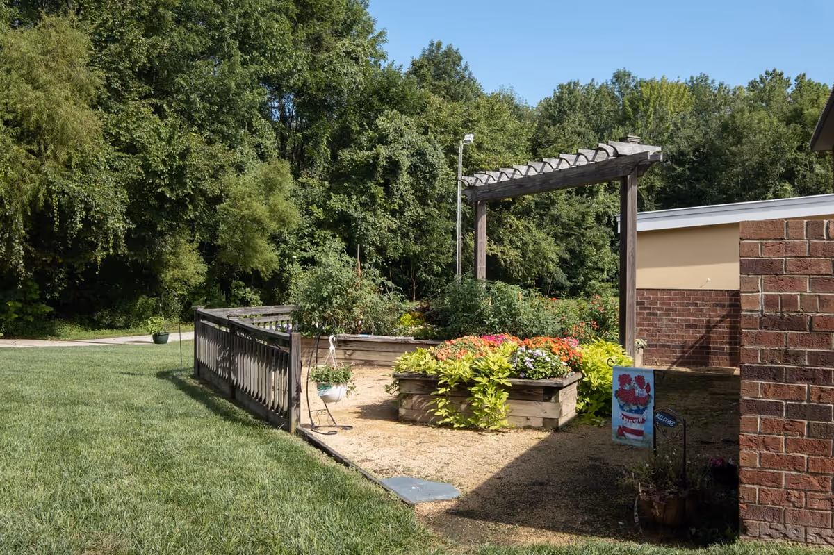A sunny outdoor garden area with raised wooden flower beds filled with colorful flowers and green plants. There is a wooden pergola structure above one of the flower beds. The garden is bordered by a wooden fence and surrounded by lush green trees and grass. A brick building is partially visible on the right side of the image.