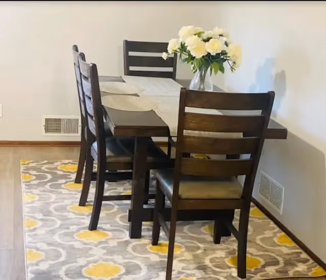 A dining area with a dark wooden table and six matching chairs placed on a patterned rug with gray, white, and yellow designs. A vase with white flowers is centered on the table. The walls are light-colored with a vent visible near the floor.