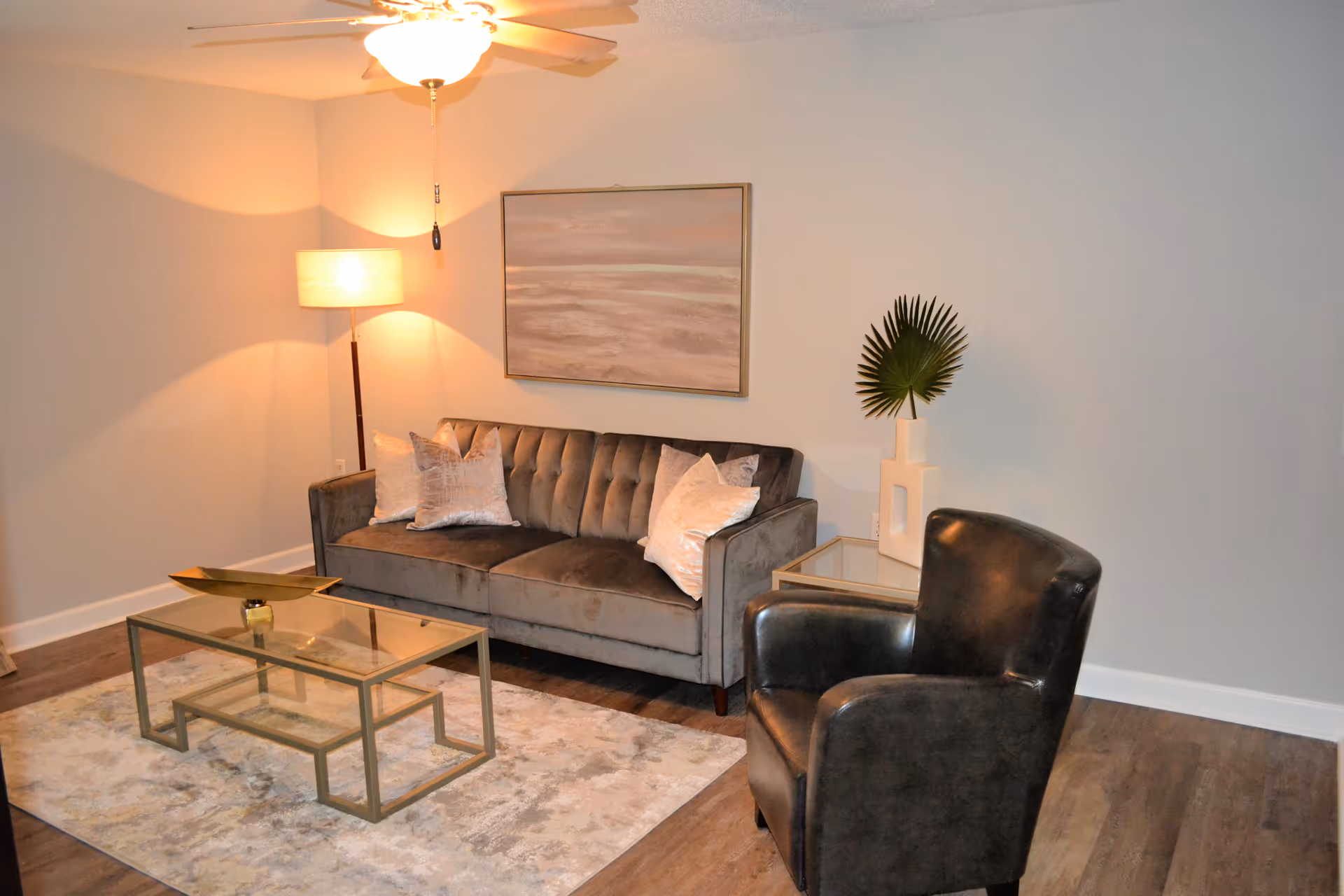 A cozy living room with a gray velvet sofa adorned with three light-colored pillows, a glass coffee table with a decorative golden tray, a black leather armchair, a floor lamp with a warm light, a framed abstract painting on the wall, and a side table with a white vase holding a green palm leaf.