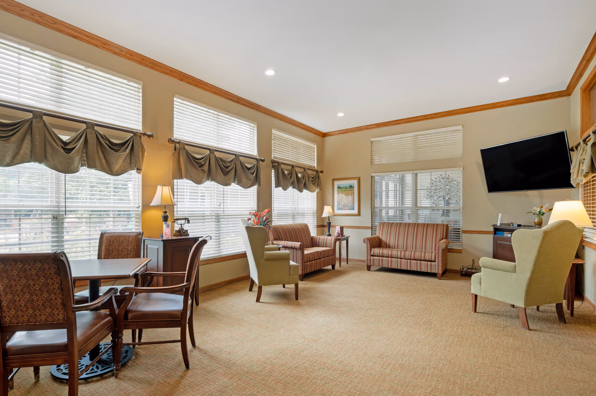 A bright and spacious living room with large windows covered by white blinds and green valances. The room features two striped sofas, two green armchairs, a wooden table with four chairs, several lamps, a wall-mounted flat-screen TV, and framed artwork on the walls. The carpet is beige, and the walls have wooden trim.