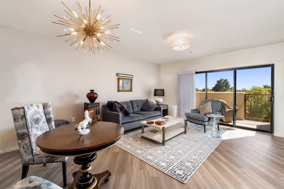 A bright living room with a gray sofa, a patterned armchair, a white and gold coffee table on a geometric rug, and a round wooden table with two floral upholstered chairs. A modern gold chandelier hangs from the ceiling. Sliding glass doors lead to a balcony with a view of trees and a clear sky.