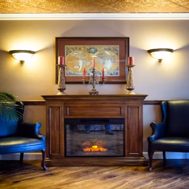 A wooden fireplace mantel with decorative candles and a framed map above, flanked by two armchairs and wall sconces.
