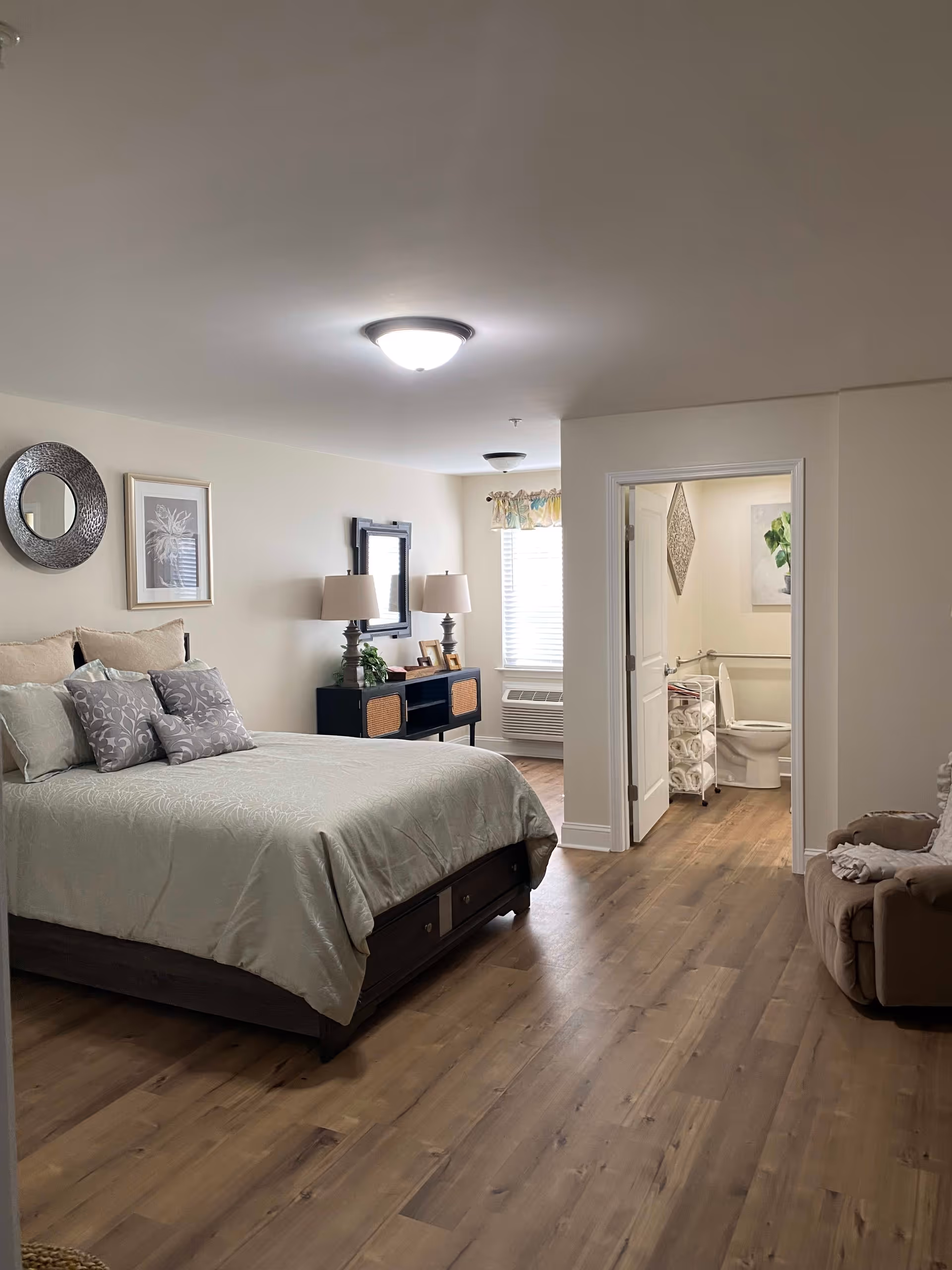 A spacious bedroom with a large bed covered in light green bedding and multiple pillows. There is a dark wooden dresser with two lamps and framed pictures on top, a round decorative mirror and framed artwork on the wall. The room has wooden flooring and a window with blinds and a floral valance. To the right, an open door reveals a bathroom with a toilet, a towel rack with rolled towels, and wall art. A beige recliner chair is partially visible on the right side of the image.
