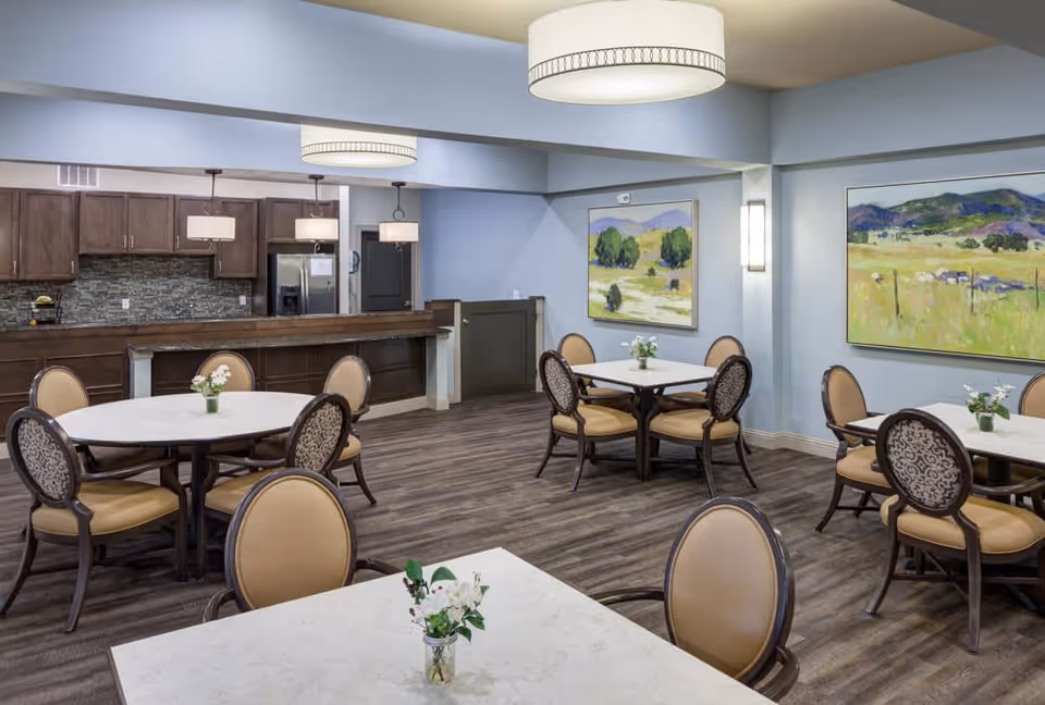 A dining room with several round and square tables, each surrounded by four cushioned chairs. The tables have small flower arrangements. The room features wood flooring, light blue walls, two large landscape paintings, and modern ceiling lights. In the background, there is a kitchen area with dark wood cabinets, a stone backsplash, and stainless steel appliances.