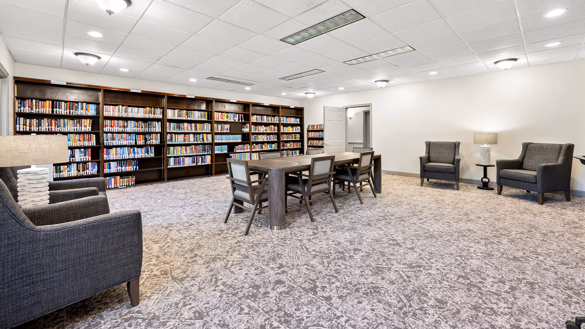 A spacious library room with multiple dark wood bookshelves filled with books along the back wall. In the center, there is a large wooden table surrounded by six chairs. The room has a patterned carpet, white walls, and several ceiling lights. There are also three gray upholstered armchairs with two small side tables and lamps near the walls.