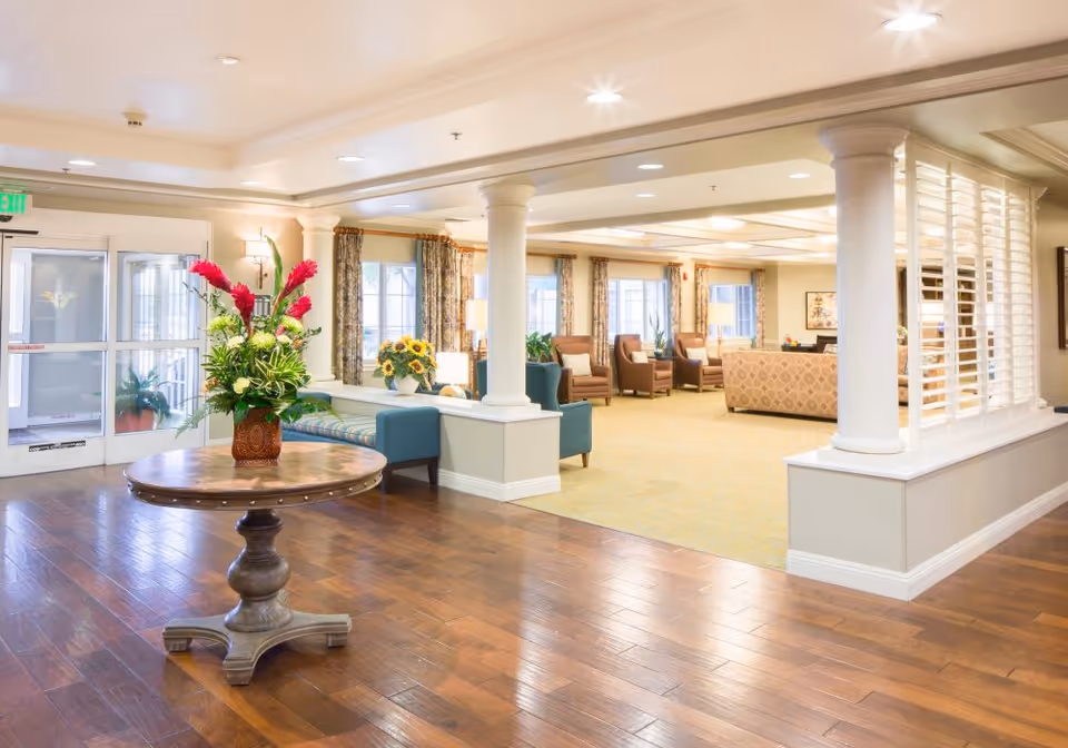 A bright and spacious senior living facility common area with wooden flooring, a round wooden table with a floral arrangement, comfortable seating including armchairs and sofas, large windows with curtains, and white columns separating the spaces.