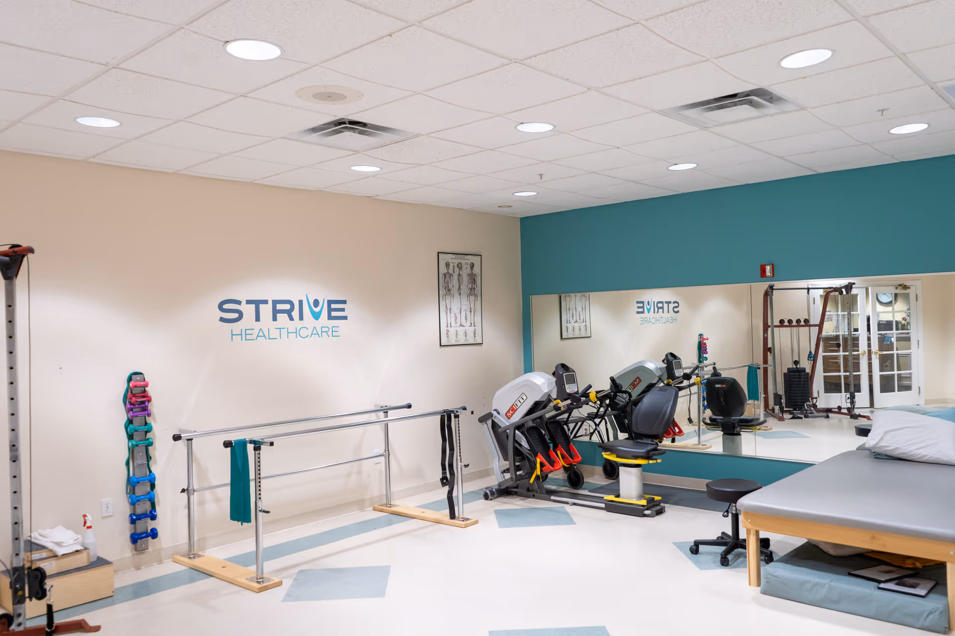 A rehabilitation therapy room with exercise equipment including a recumbent bike, parallel bars for walking practice, a padded therapy table with a pillow, and a large mirror on one wall. The walls are painted beige and teal, with the logo 'STRIVE HEALTHCARE' displayed on one wall. There are anatomical charts and various small weights organized on a rack.