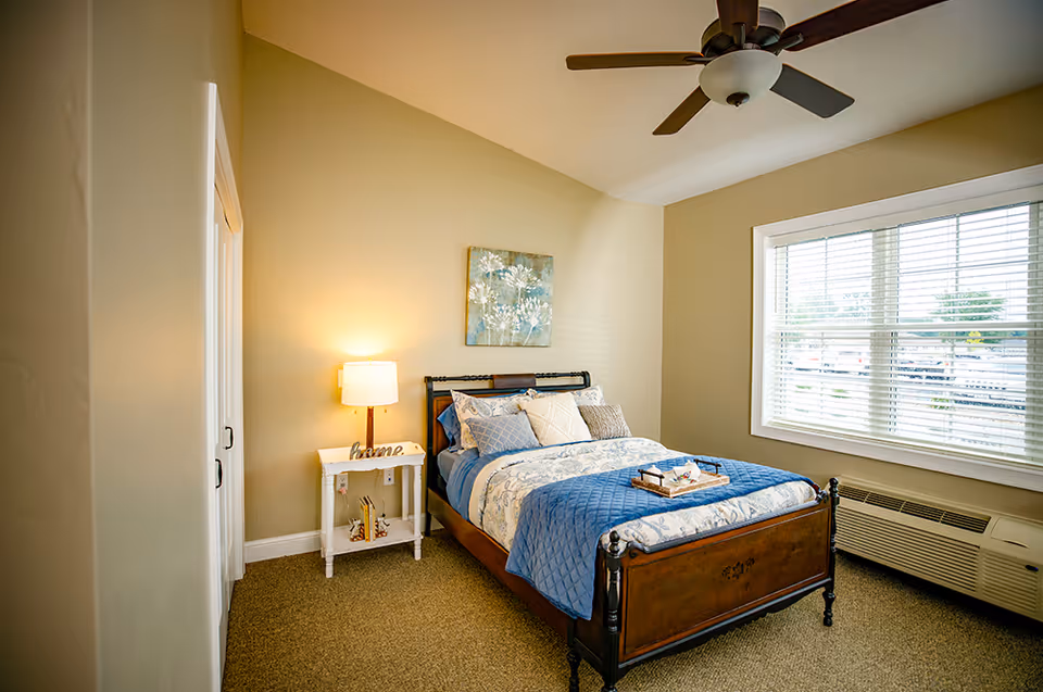 A cozy bedroom with a wooden bed frame, blue and white bedding, and multiple pillows. A small white nightstand with a lamp and decorative items is beside the bed. A large window with blinds lets in natural light, and a ceiling fan is mounted on the ceiling. The walls are painted beige, and there is a piece of floral artwork hanging above the bed.
