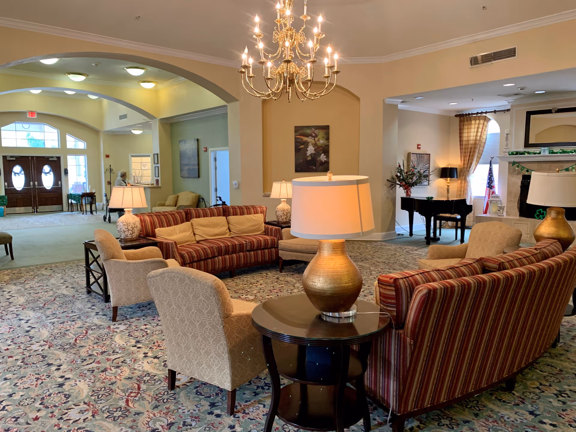 A spacious and well-lit senior living facility common area with patterned carpet, striped sofas with beige cushions, beige armchairs, and several table lamps. A grand chandelier hangs from the ceiling. In the background, there is a black grand piano near a fireplace decorated with green garlands and an American flag. The entrance doors with glass panels are visible in the distance, along with a person standing near the reception area.