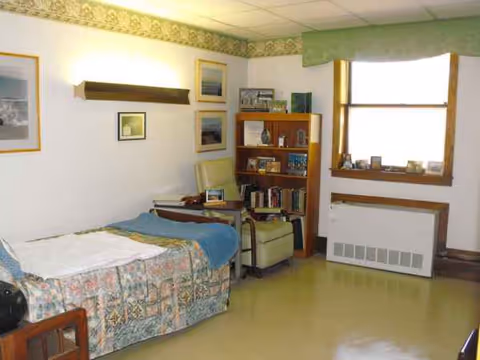 A small nursing home bedroom with quilted bed, chair, bookshelf, and window.