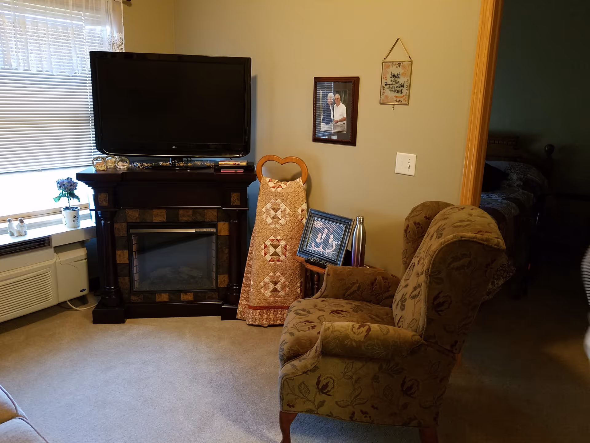 A cozy living room area with a floral patterned armchair facing a black electric fireplace with a flat-screen TV mounted above it. To the left, there is a window with blinds and a small potted plant on the windowsill. A quilted decorative piece is draped over a wooden chair next to the fireplace. On the wall, there is a framed photo of two people and a hanging decorative sign. A small side table holds a framed picture and a metallic water bottle. An open doorway leads to a dimly lit bedroom with a bed visible.