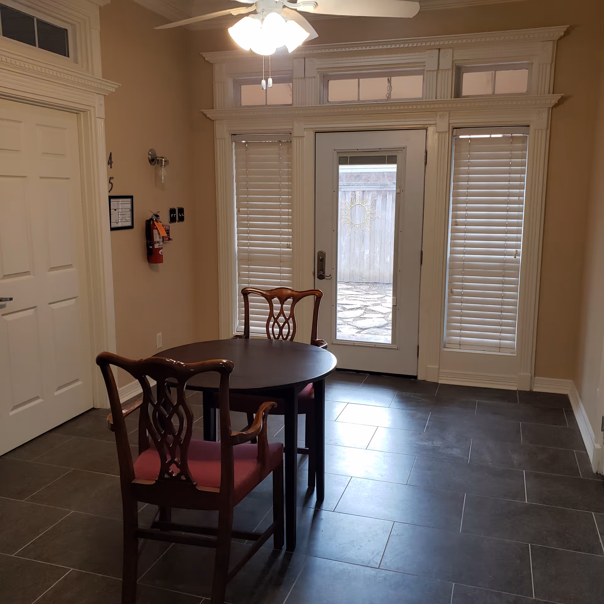 A small interior room with a round wooden table and two wooden chairs with red cushions. The room has beige walls, dark tiled flooring, and a white door with glass panels leading outside. There are windows with blinds on either side of the door and a ceiling fan with lights above the table. A fire extinguisher is mounted on the wall to the left.