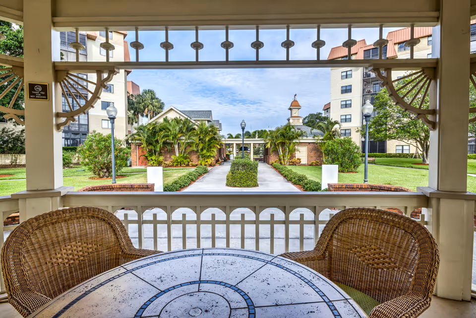 View from a covered porch with a round table and two wicker chairs looking out onto a landscaped pathway lined with palm trees and bushes, flanked by multi-story buildings under a partly cloudy sky.